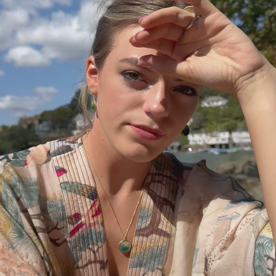 A women wears matching teal ceramic and glass jewellery with gold detailing, set against a seaside background. She wears two  rings, a round pendant necklace and oval drop earrings. 