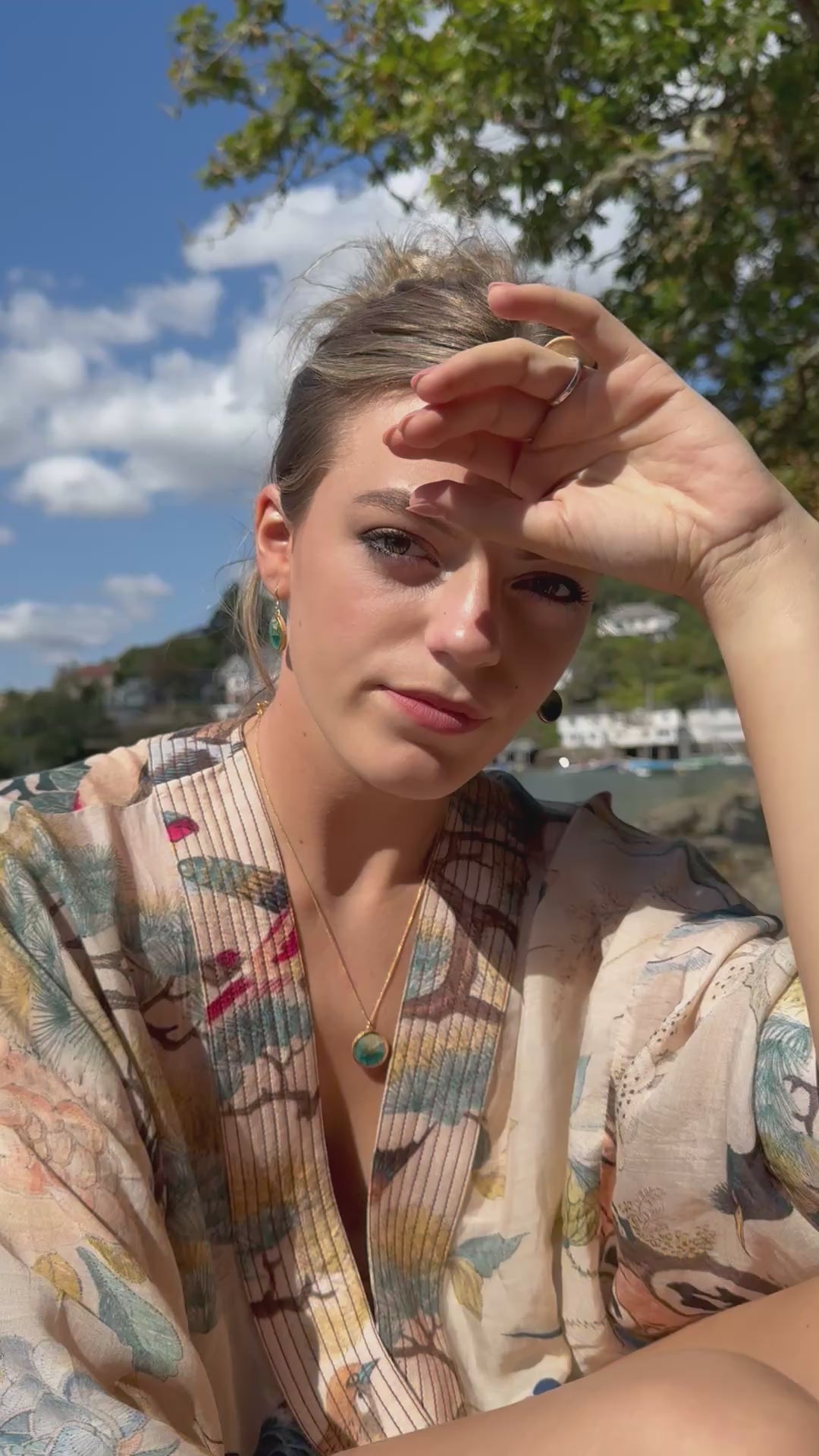 A women wears matching teal ceramic and glass jewellery with gold detailing, set against a seaside background. She wears two  rings, a round pendant necklace and oval drop earrings. 