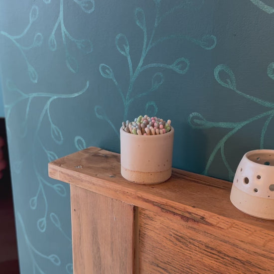 match striker pot on fireplace is picked up, a match taken and struck on the striker pad at base, a handmade votive is then lit with the match. Set on a rustic wood fireplace.