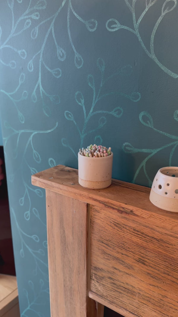 match striker pot on fireplace is picked up, a match taken and struck on the striker pad at base, a handmade votive is then lit with the match. Set on a rustic wood fireplace.