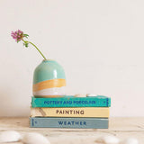 A mini shoreline vase with a pink wildflower sits on top of a stack of blue and cream vintage books with shells in the foreground.