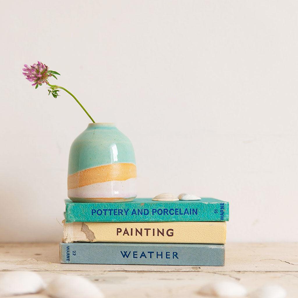A mini shoreline vase with a pink wildflower sits on top of a stack of blue and cream vintage books with shells in the foreground.