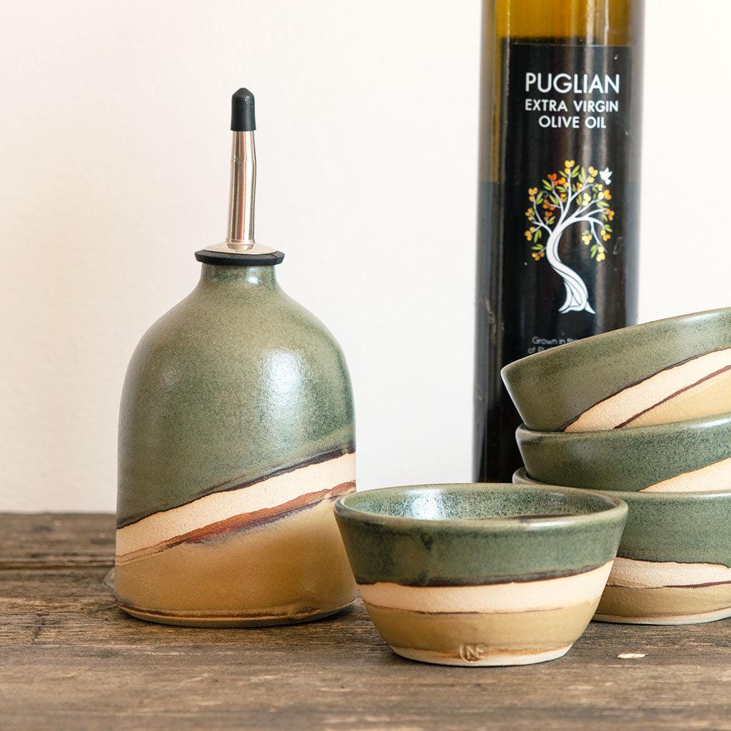 Oil pourer in a satin green glaze and light mustard glaze separated by a band of raw exposed clay. The oil pourer is next to a stack of matching mini bowls and a bottle of olive oil