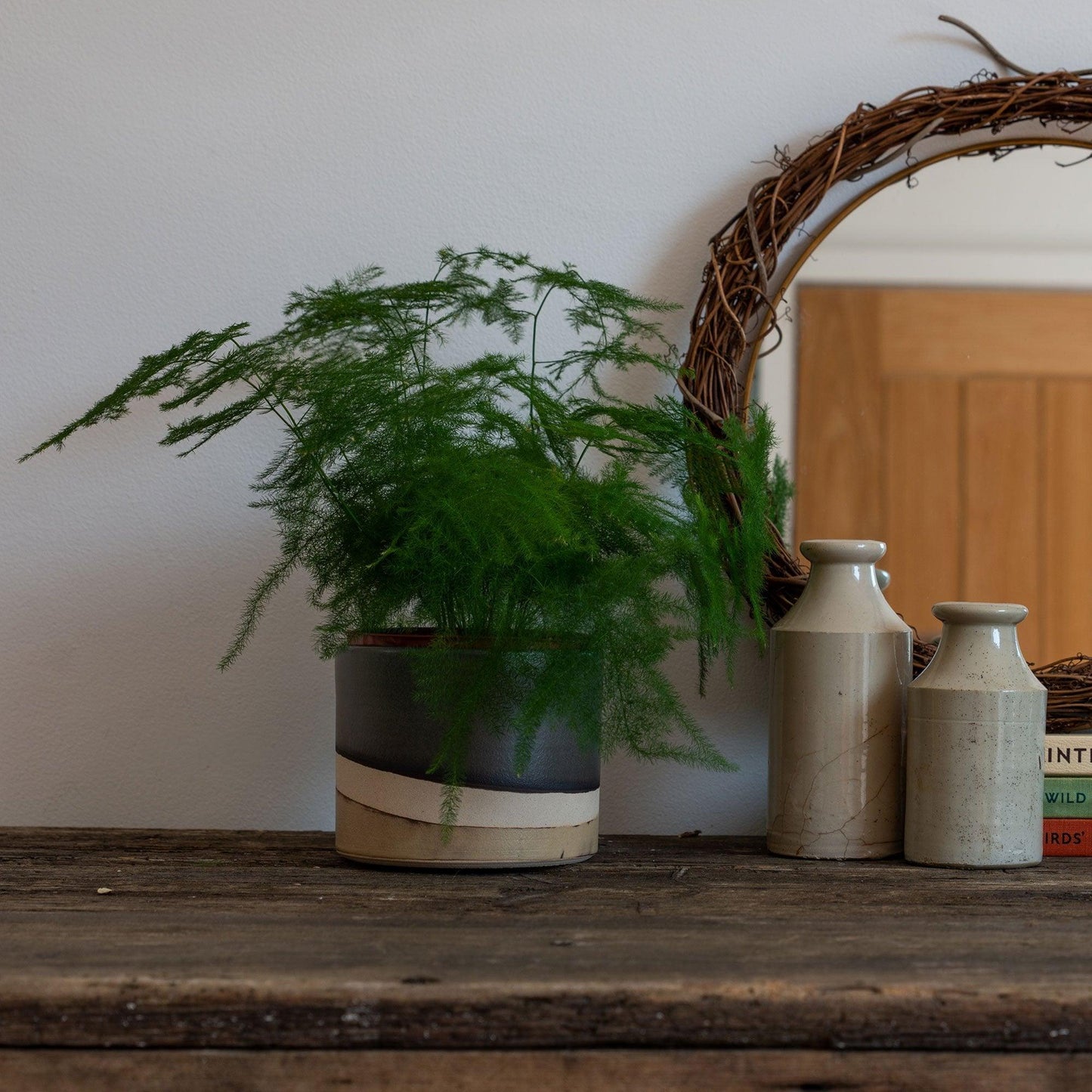 A large moorland plant pot containing a large fern plant on a dark wooden table next to stoneware vases, a wreath and a circular mirror