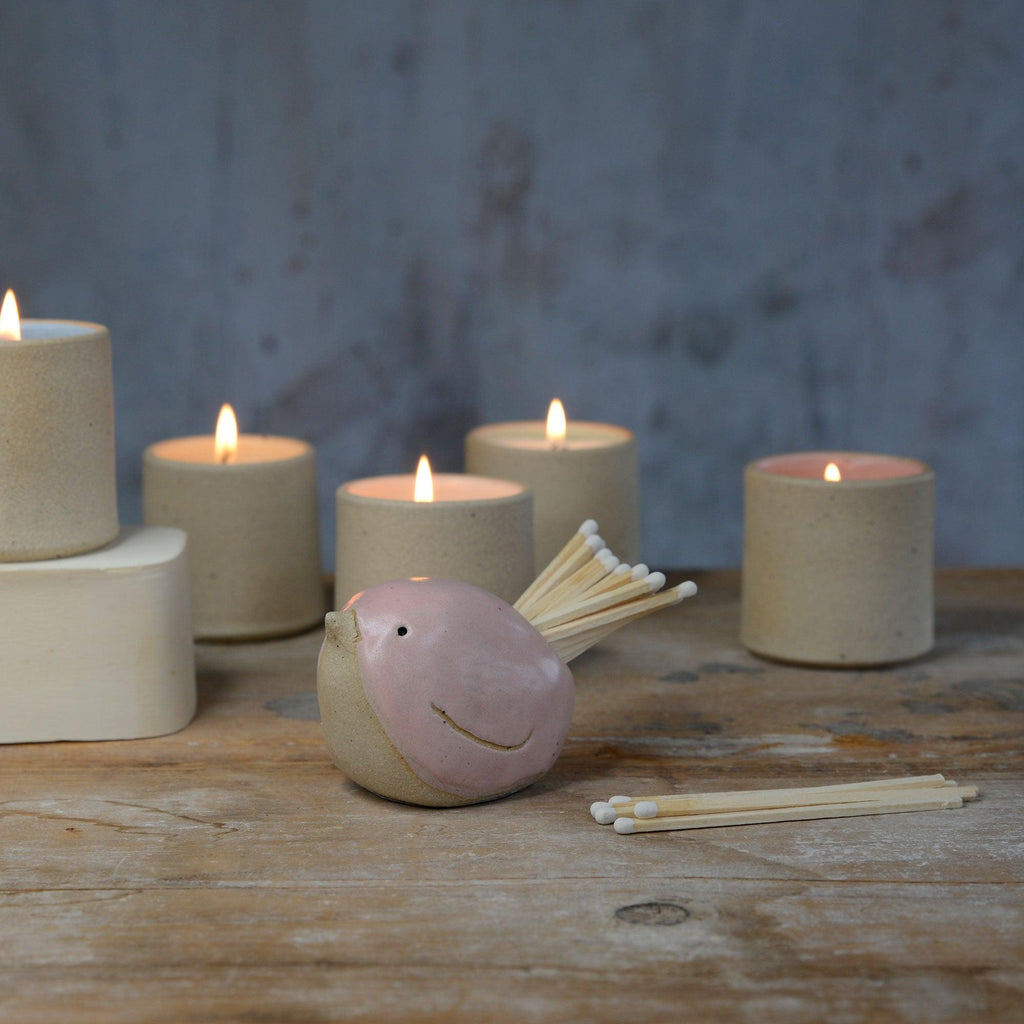 Pink bird pot filled with white matches in front of lit stoneware candles