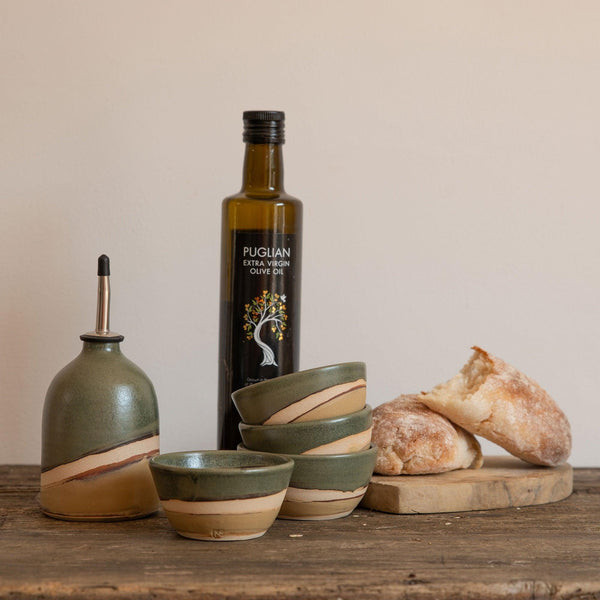 Moorland oil pourer next to a stack of matching mini bowls, an olive oil bottle and some bread on a wooden board