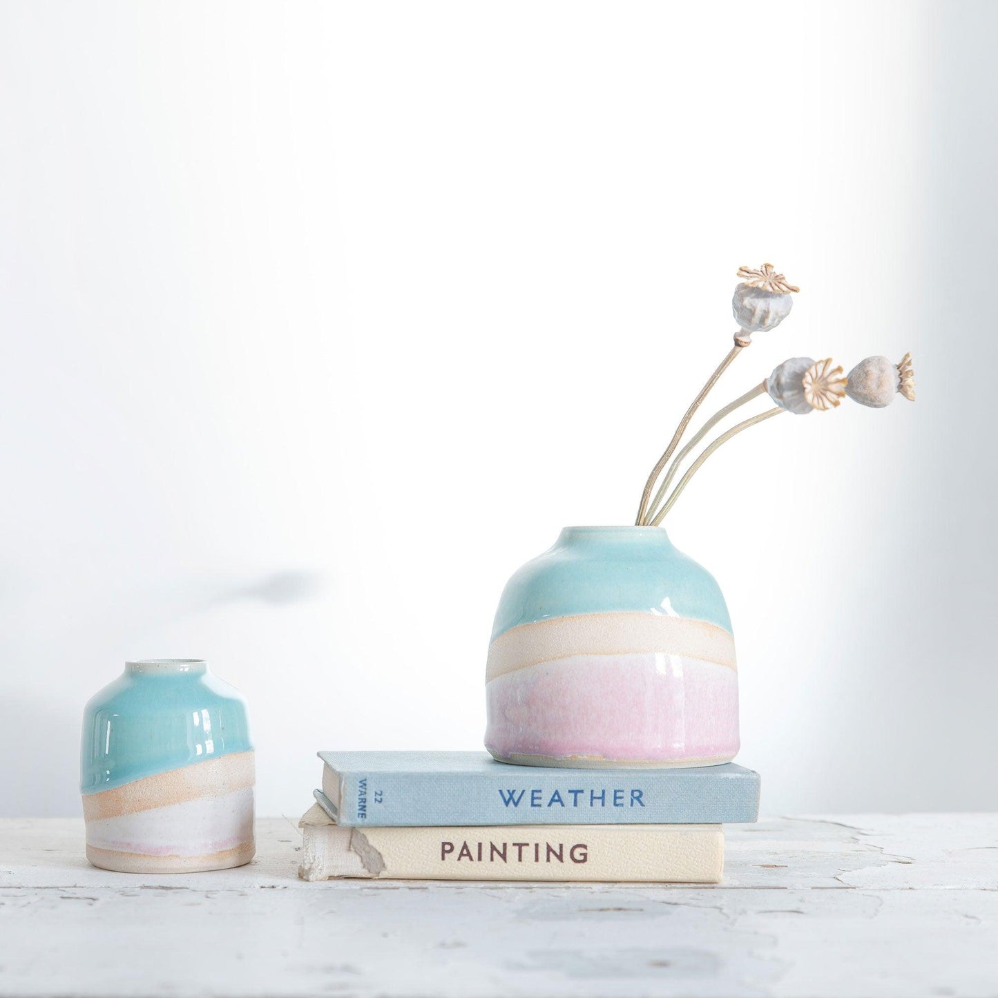 Shoreline dome vase filled with three dried poppy head stems on a couple of pastel vintage books, a mini shoreline vase sits to the left.