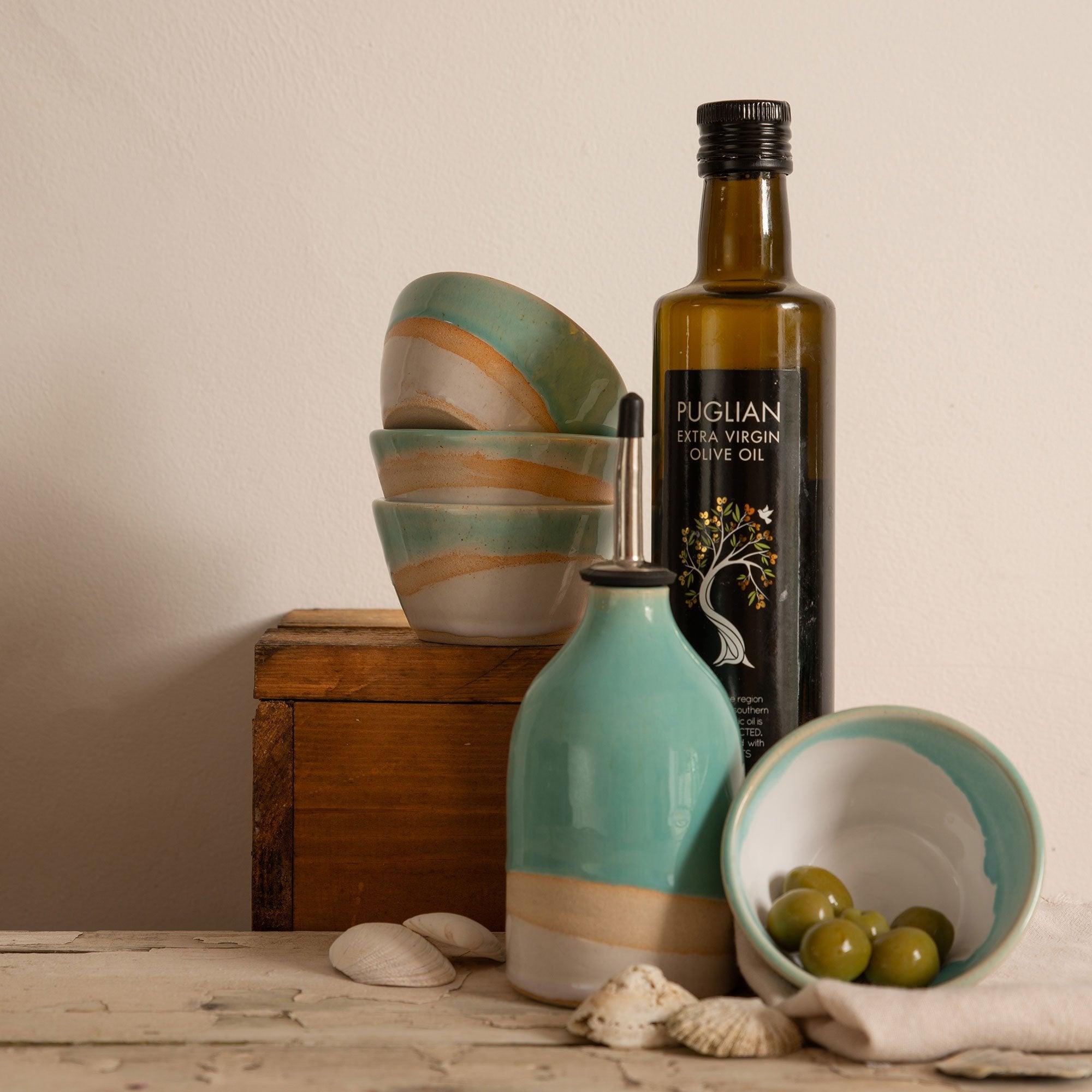 Oil pourer amidst matching mini bowl of olives, a stack of mini bowls in the rear and a bottle of olive oil