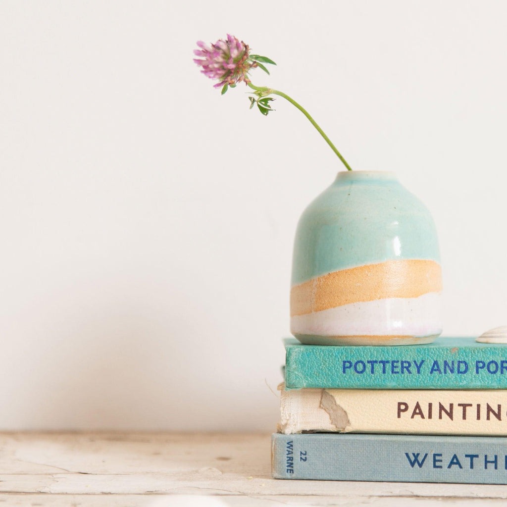 A mini Shoreline vase in glossy turquoise and pale pink glazes with exposed burnt-orange clay on top of a stack of blue and cream vintage books. A single pink wildflower is in the vase.