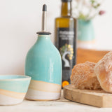 Shoreline oil pourer in shiny turquoise glaze and pale pink glaze separated by a band of raw exposed clay. The pourer is next to some bread and a matching mini bowl with an olive oil bottle in the background