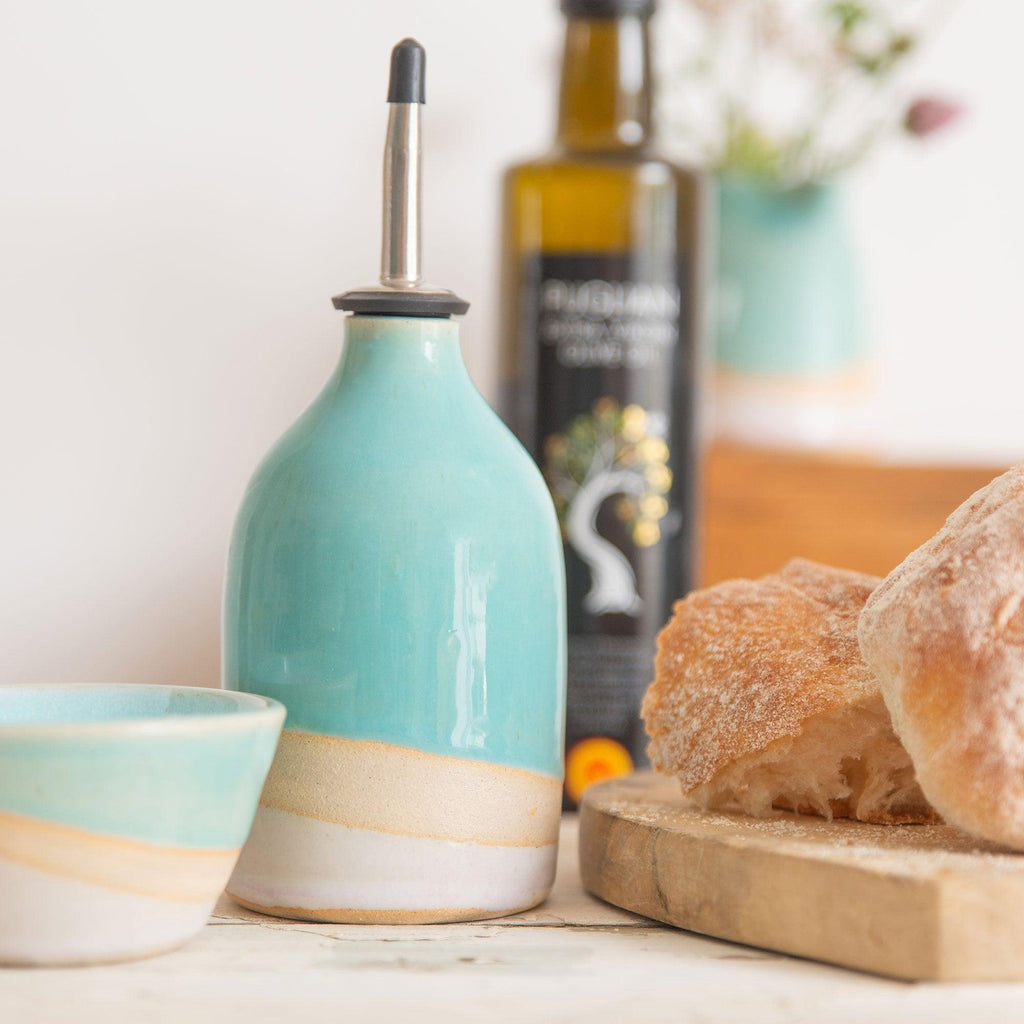 Shoreline oil pourer in shiny turquoise glaze and pale pink glaze separated by a band of raw exposed clay. The pourer is next to some bread and a matching mini bowl with an olive oil bottle in the background