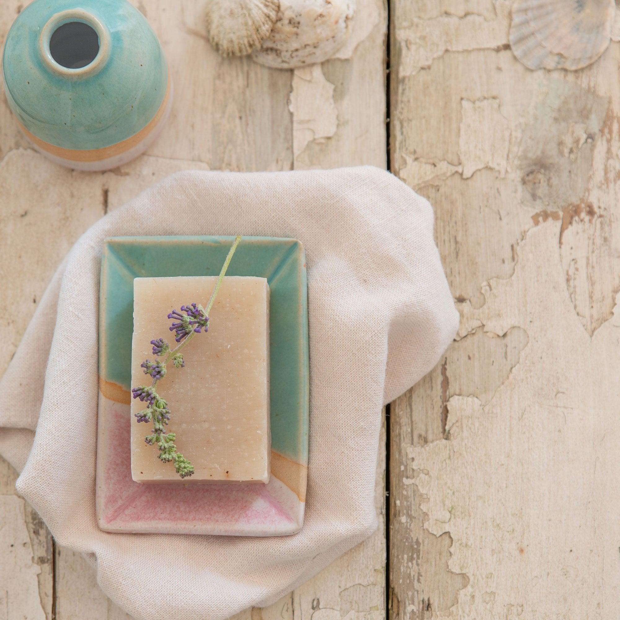 Purple wildflower sprig on top of a bar of soap in the shoreline soap dish. A matching Shoreline vase is next to it in the top left