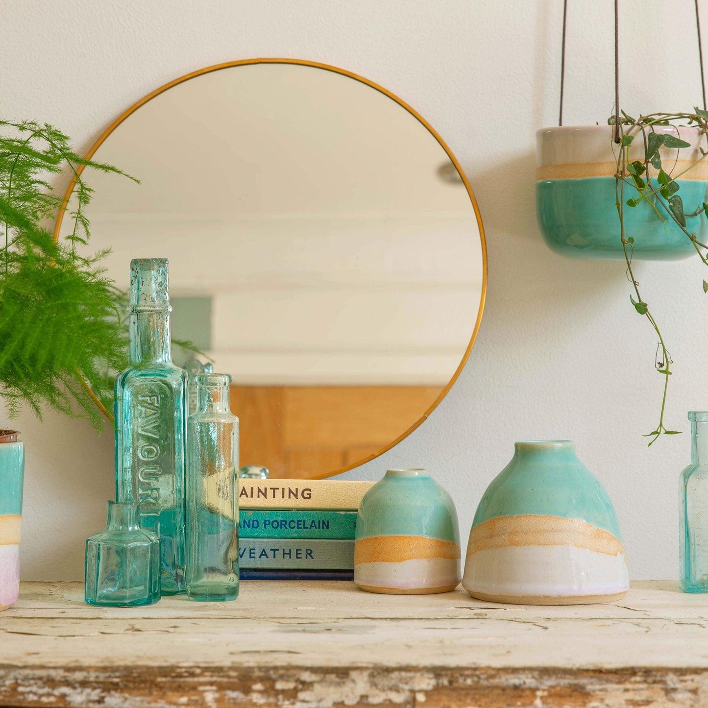 A shoreline hanging planter hangs to the right above an aged wooden mantle piece decorated with a mini and dome shoreline vase, a stack of vintage books and a cluster of turquoise glass bottles in front of a circular mirror.