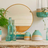 A shoreline hanging planter hangs to the right above an aged wooden mantle piece decorated with a mini and dome shoreline vase, a stack of vintage books and a cluster of turquoise glass bottles in front of a circular mirror.