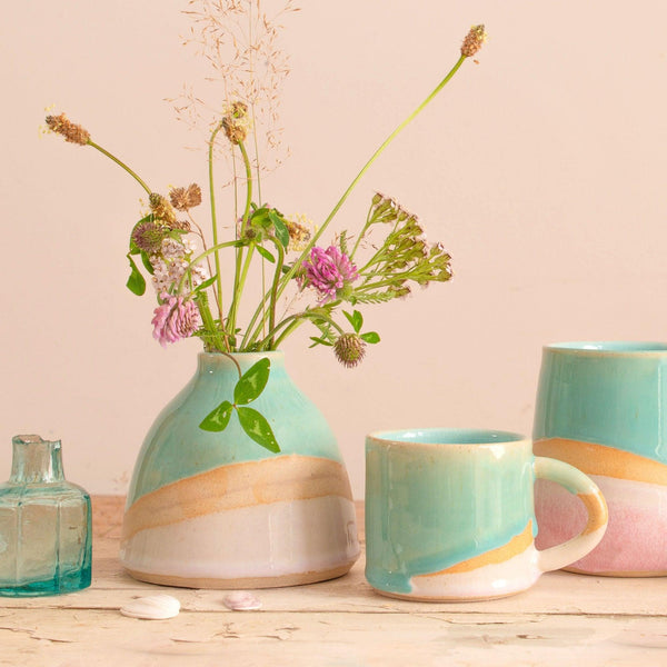 Shoreline dome vase filled with pink and green wildflowers next to a shoreline espresso mug, tumbler and a mini turquoise glass bottle.