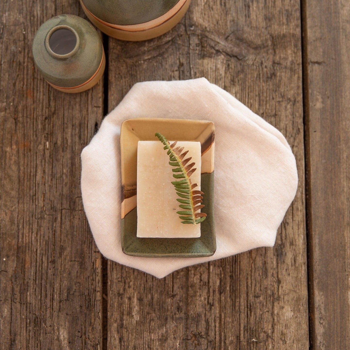A sprig of fern on top of a soap bar in the Moorland soap dish. The dish rests on top of a linen cloth next to a couple of moorland vases