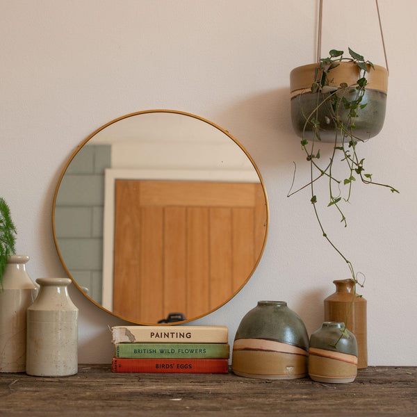 A Moorland hanging planter hanging from the top right corner holding a draping string of hearts. The planter is above a dark wooden mantle with a Moorland Dome and Mini vase, vintage books, brown ceramic vases and a circular mirror.