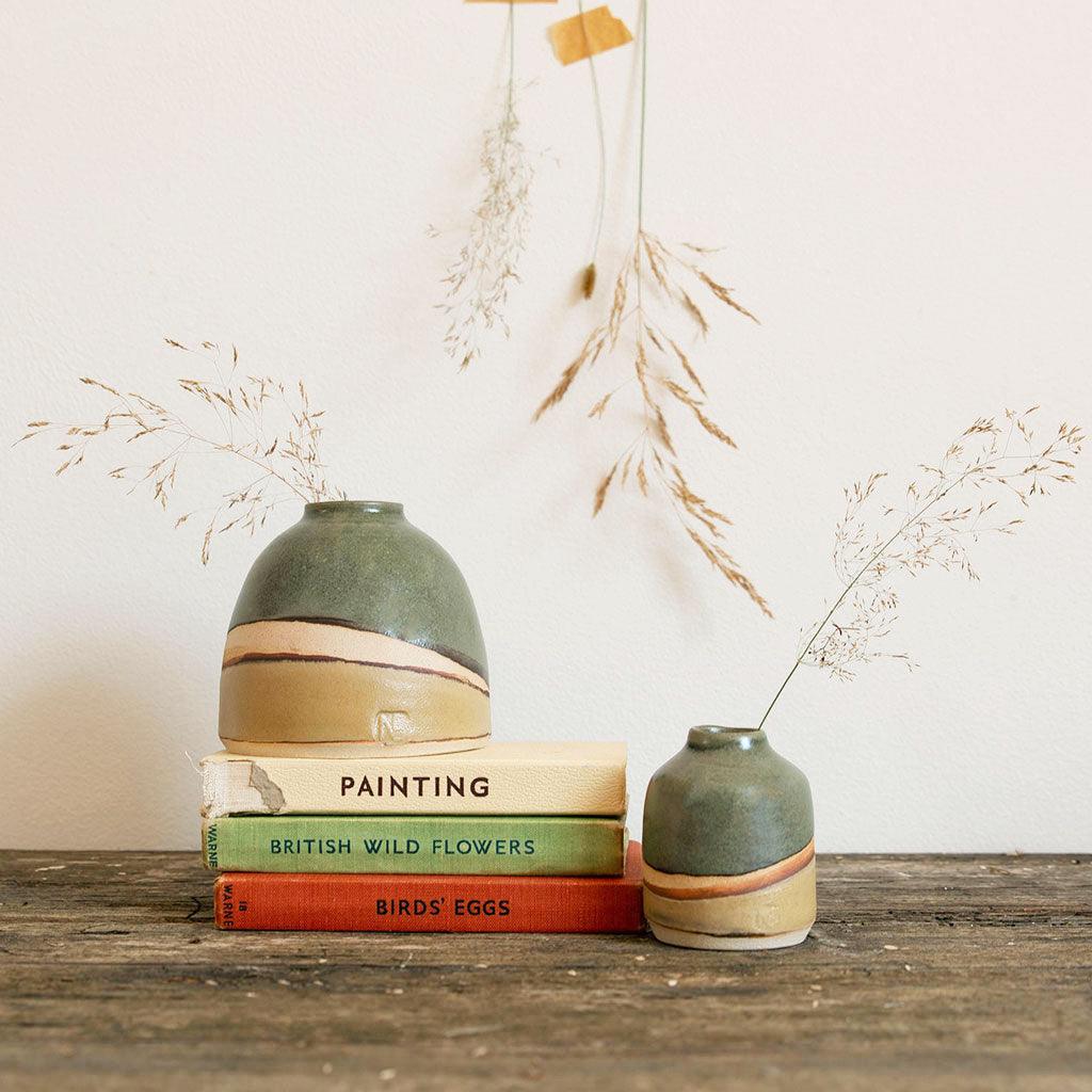 Moorland dome vase on a stack of vintage books, next to a mini moorland vase. Dried seed stems are in both vases and taped to the wall behind. The vases are glazed with a rich dark green and light brown with exposed clay in between.