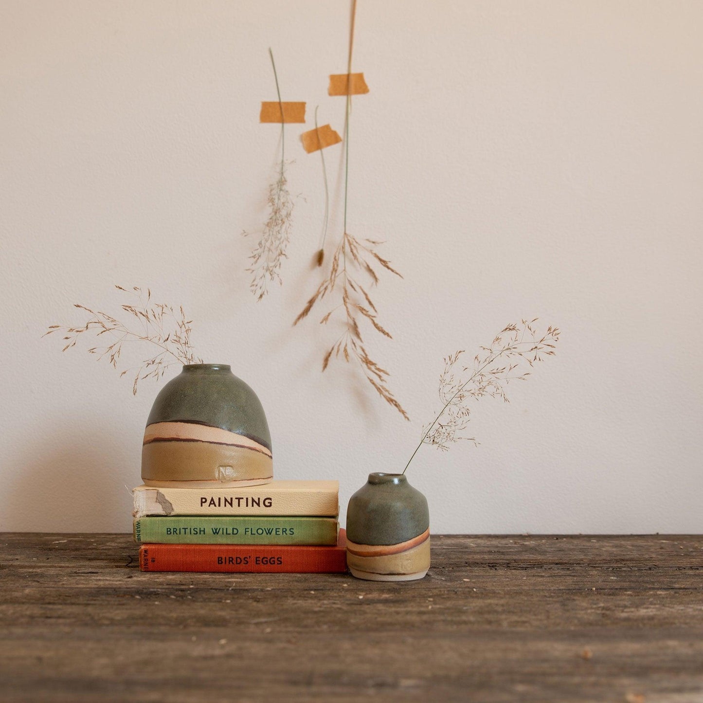 Moorland dome vase on stack of vintage books with a mini moorland vase to the right. Both vases have dried seed stems in, with similar dried stems taped to the wall behind.